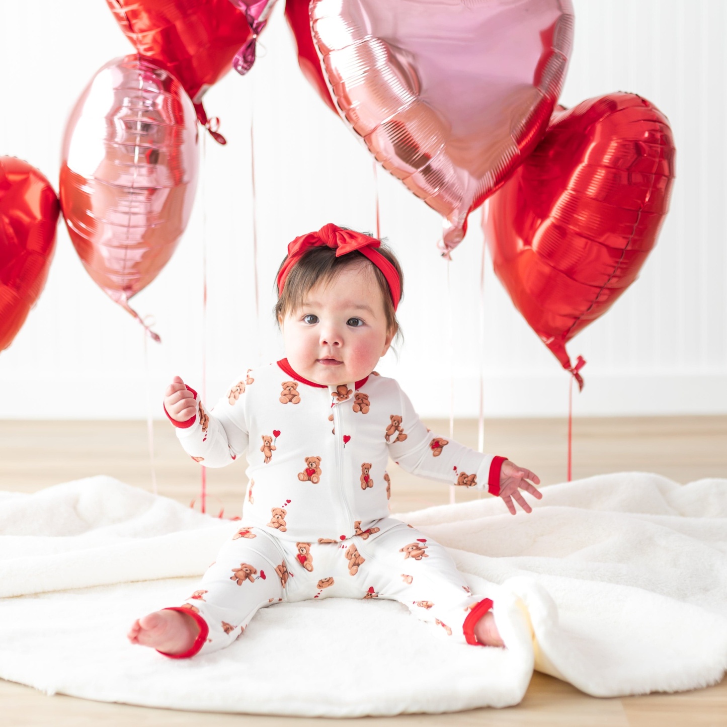 Infant sitting on a white fluffy blanket wearing the Zippered Romper in Bear Hearts with red and pink heart balloons around her