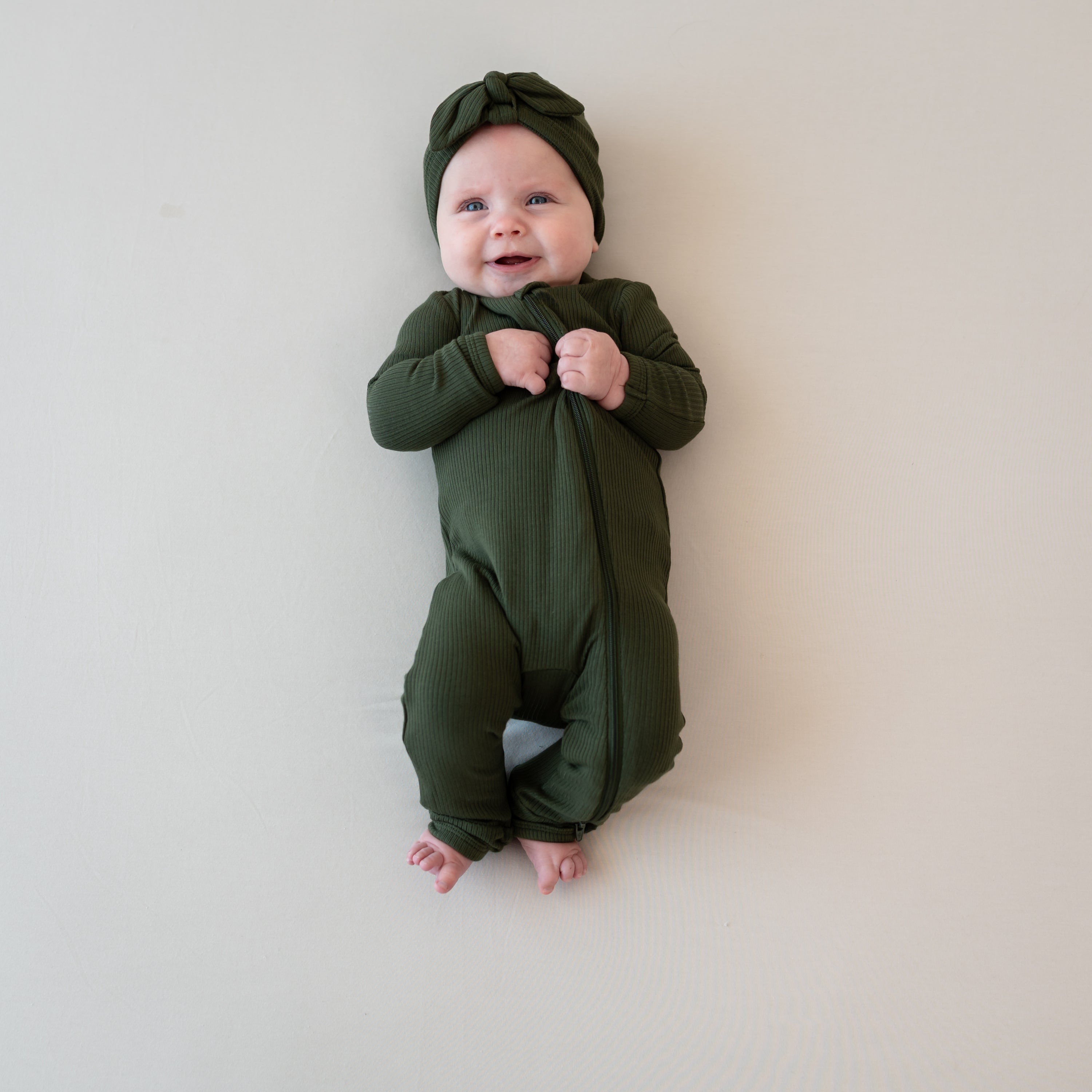 Smiling infant laying down wearing the Ribbed Zipper Romper in Fir and matching ribbed headwrap laying on a Bisque Blanket