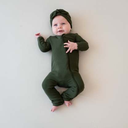 Infant laying down with one hand on her chest and the other by her shoulder wearing the Ribbed Zipper Romper in Fir and matching ribbed headwrap on a Bisque blanket