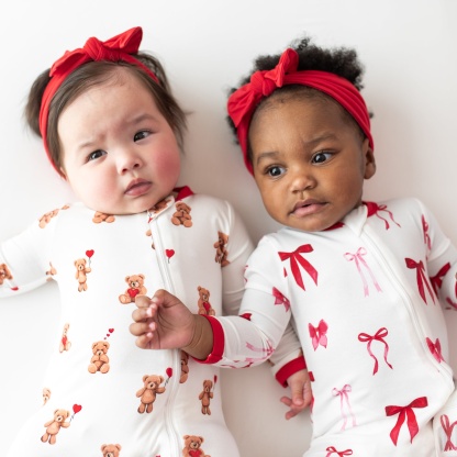 Two toddlers laying side by side wearing the Zippered Romper in Bear Hearts and Small love bow with knotted bow headbands in cardinal