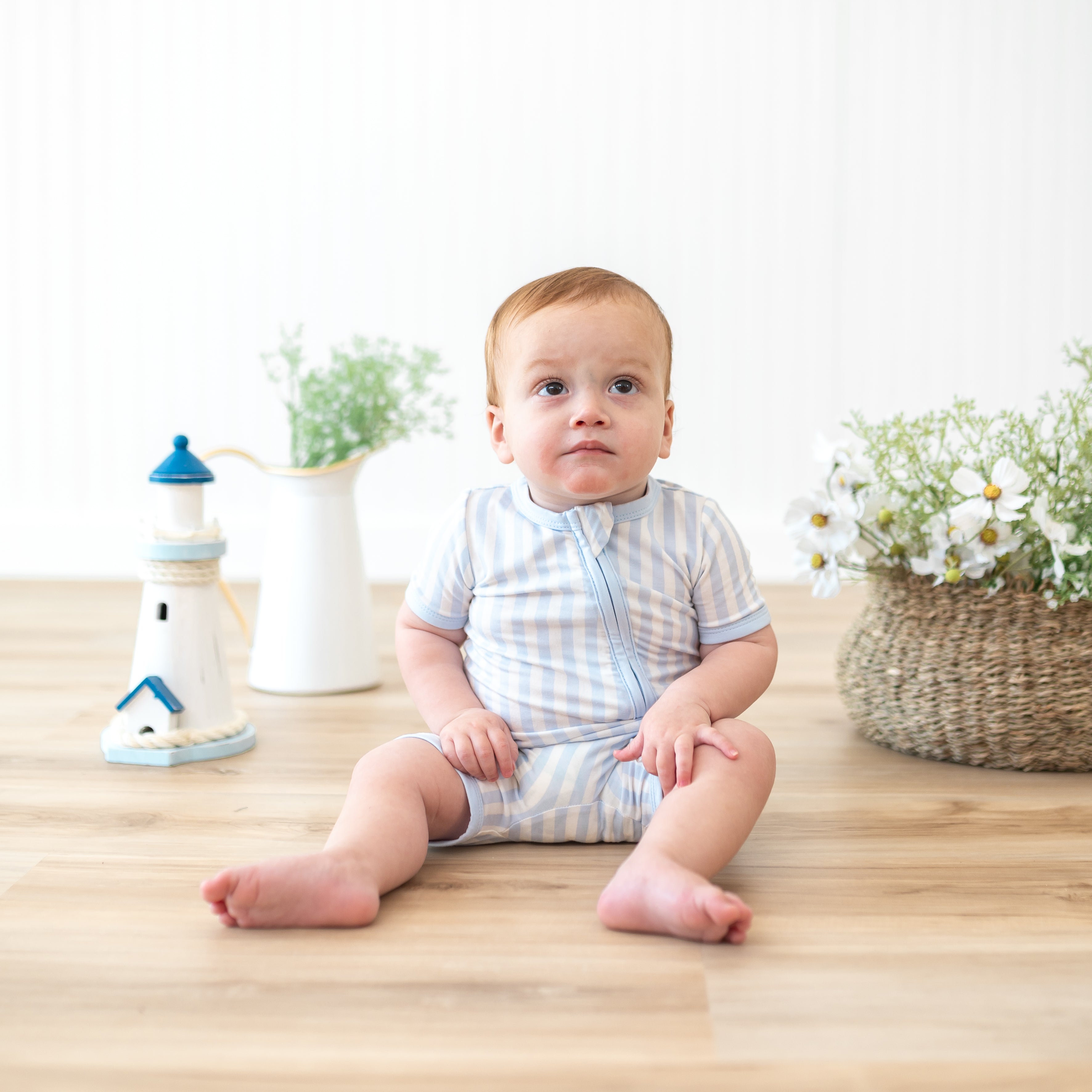 Infant sitting in front of lighthouse prop and potted flowers and plants wearing the Zipper Shortall Romper in Small Mist Stripe