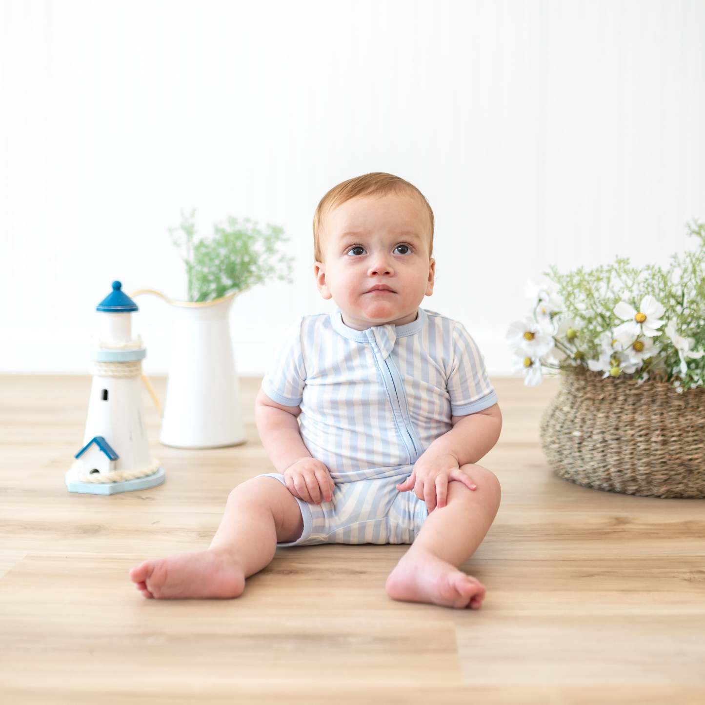 Infant sitting in front of lighthouse prop and potted flowers and plants wearing the Zipper Shortall Romper in Small Mist Stripe