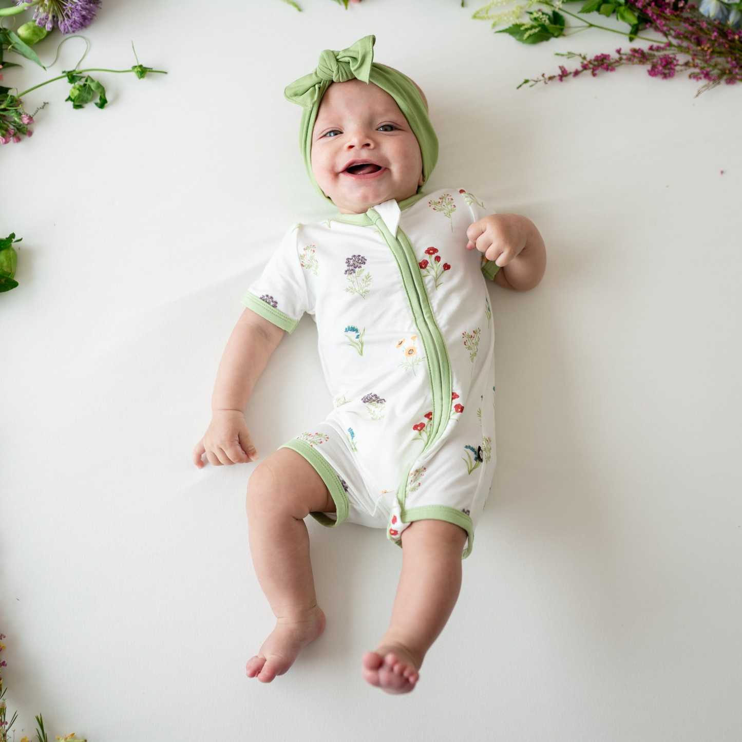 Baby smiling wearing Zippered Shortall Romper in Wildflower and Honu bow
