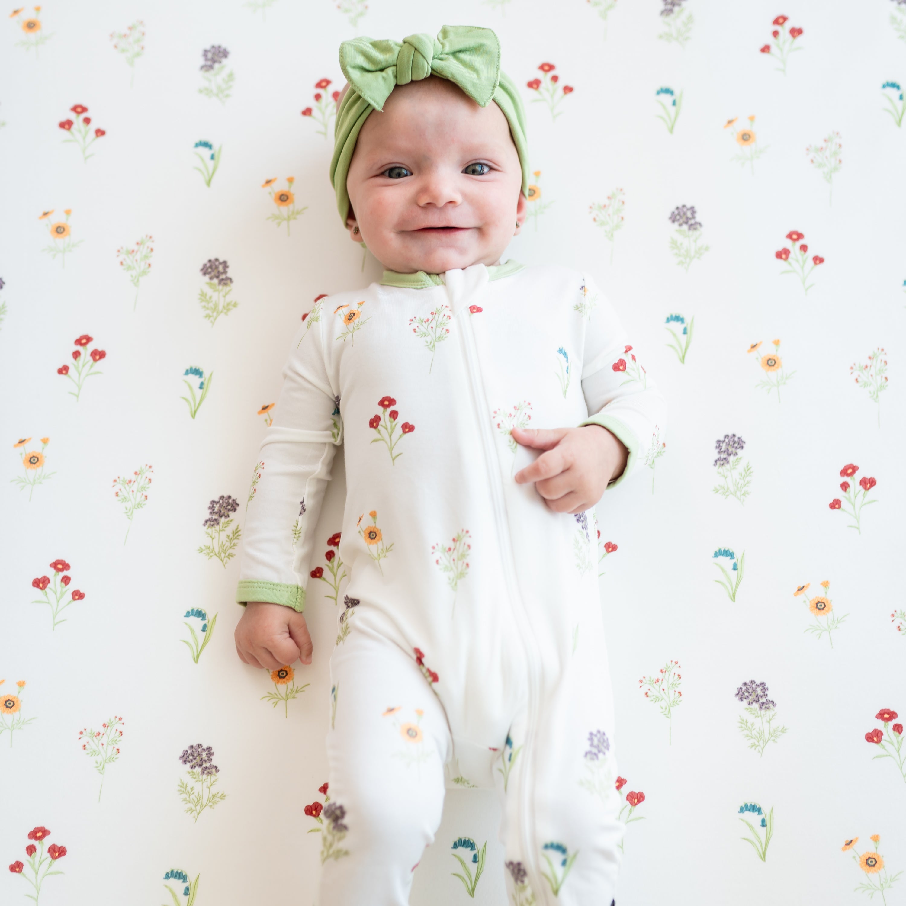 Smiling infant wearing the Zippered Romper in Wildflower with a Honu Knotted Bow Headband laying on a Wildflower crib sheet