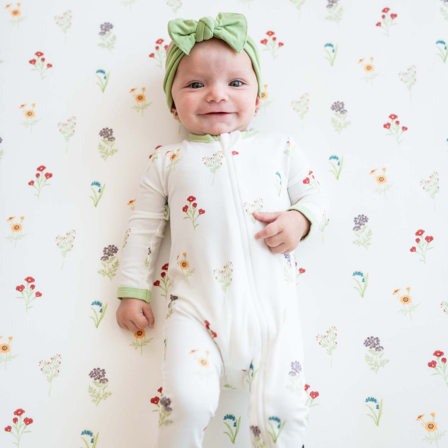 Smiling infant wearing the Zippered Romper in Wildflower with a Honu Knotted Bow Headband laying on a Wildflower crib sheet