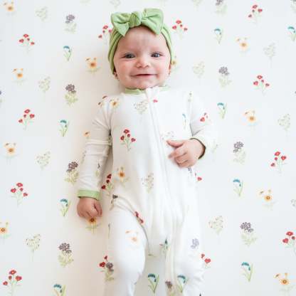 Smiling infant wearing the Zippered Romper in Wildflower with a Honu Knotted Bow Headband laying on a Wildflower crib sheet