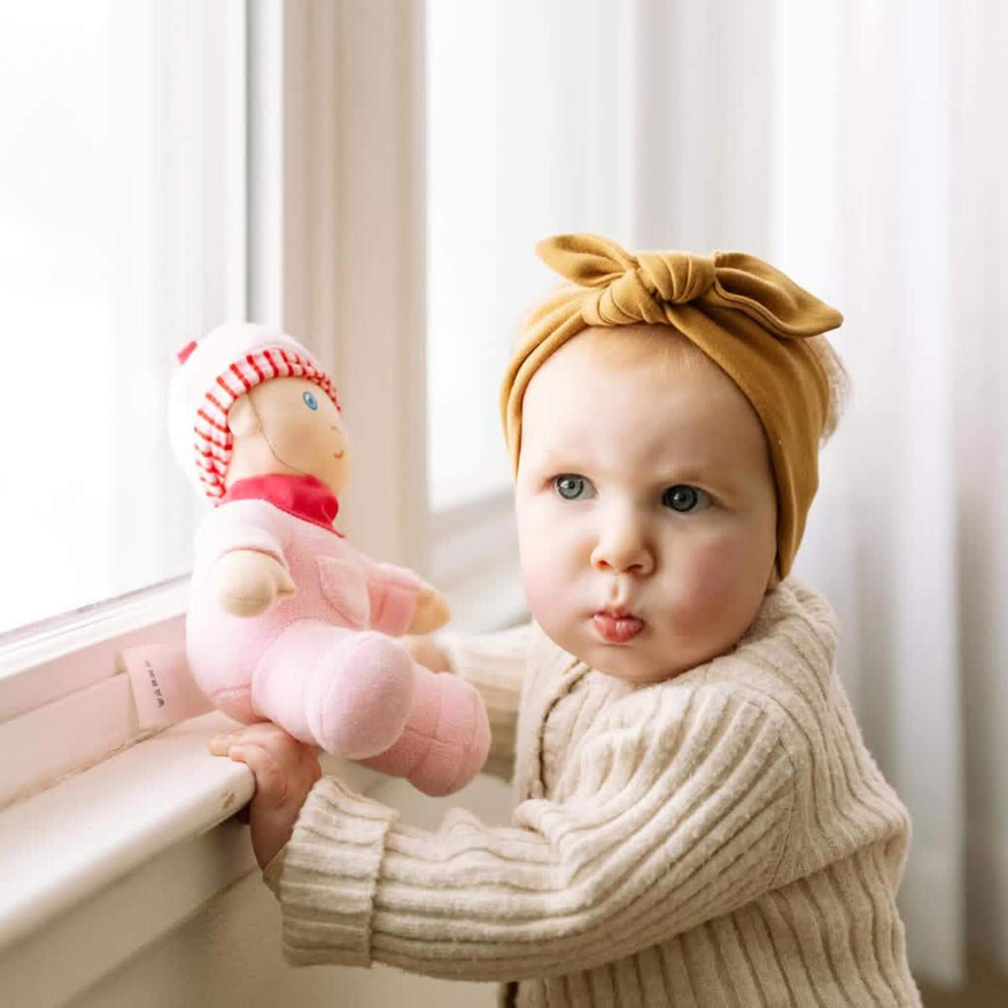 Baby holding a pink doll names Luisa with a bow headband in a softly lit room.