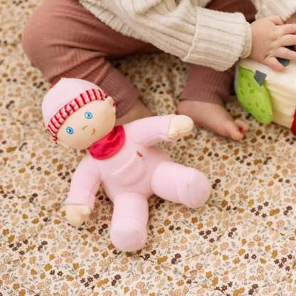 Pink plush toy with a red hat and scarf on a floral-patterned surface with a child's legs in the background.