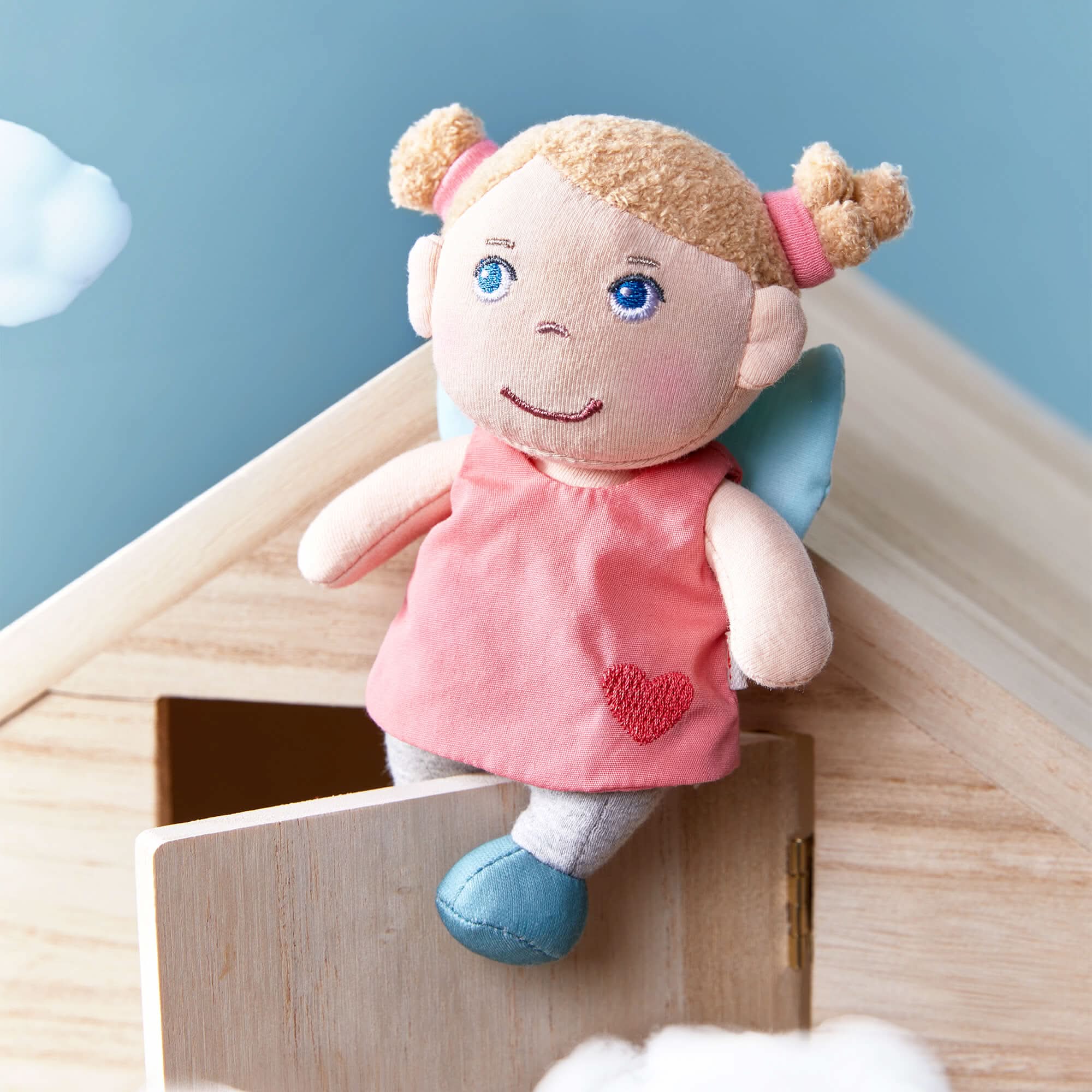 Guardian Angel Lucy doll with blonde pigtails and blue wings is sitting on a wooden structure, wearing a pink dress with a red heart.