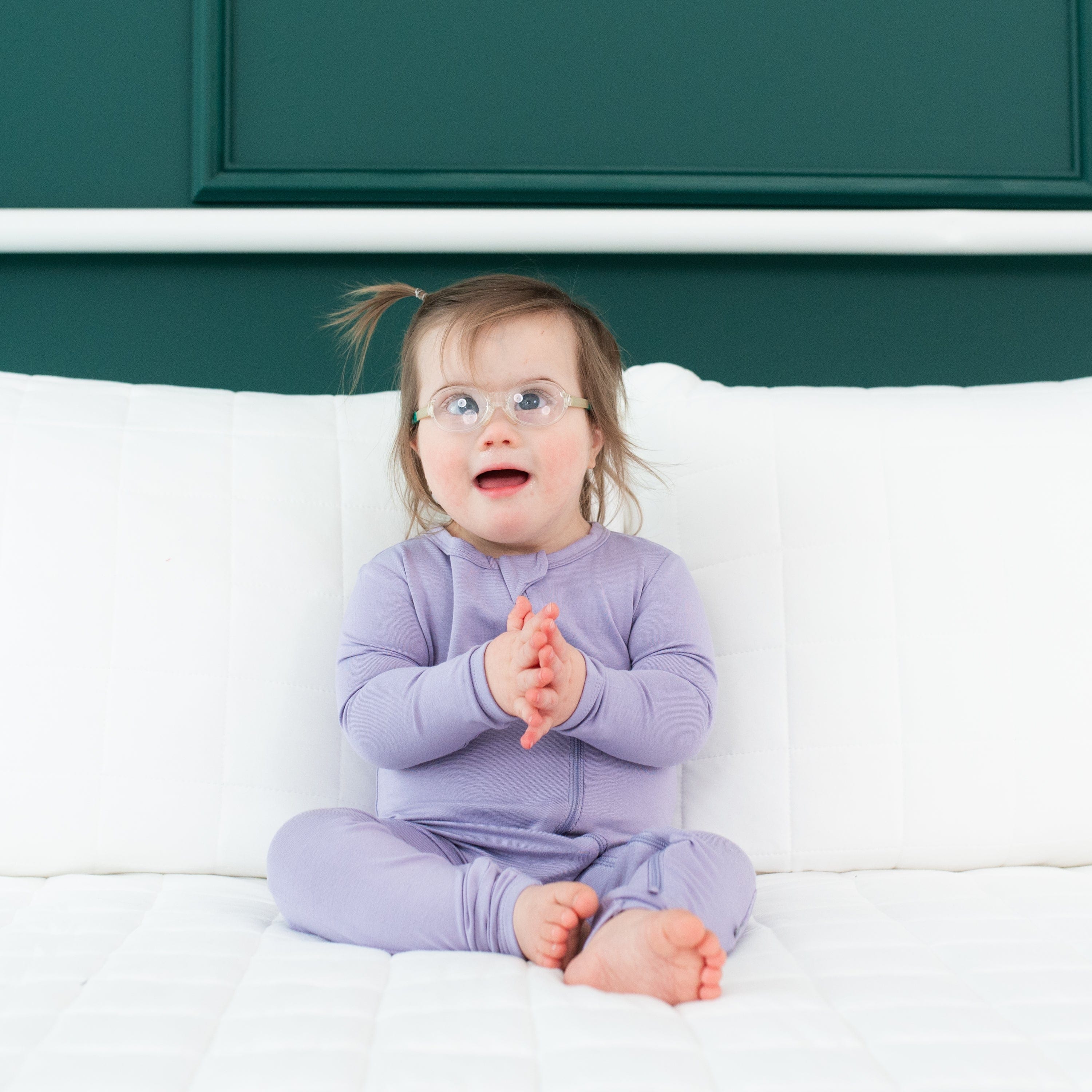Toddler sitting on a white bed infront of a green wall while wearing a Zippered Romper in Taro
