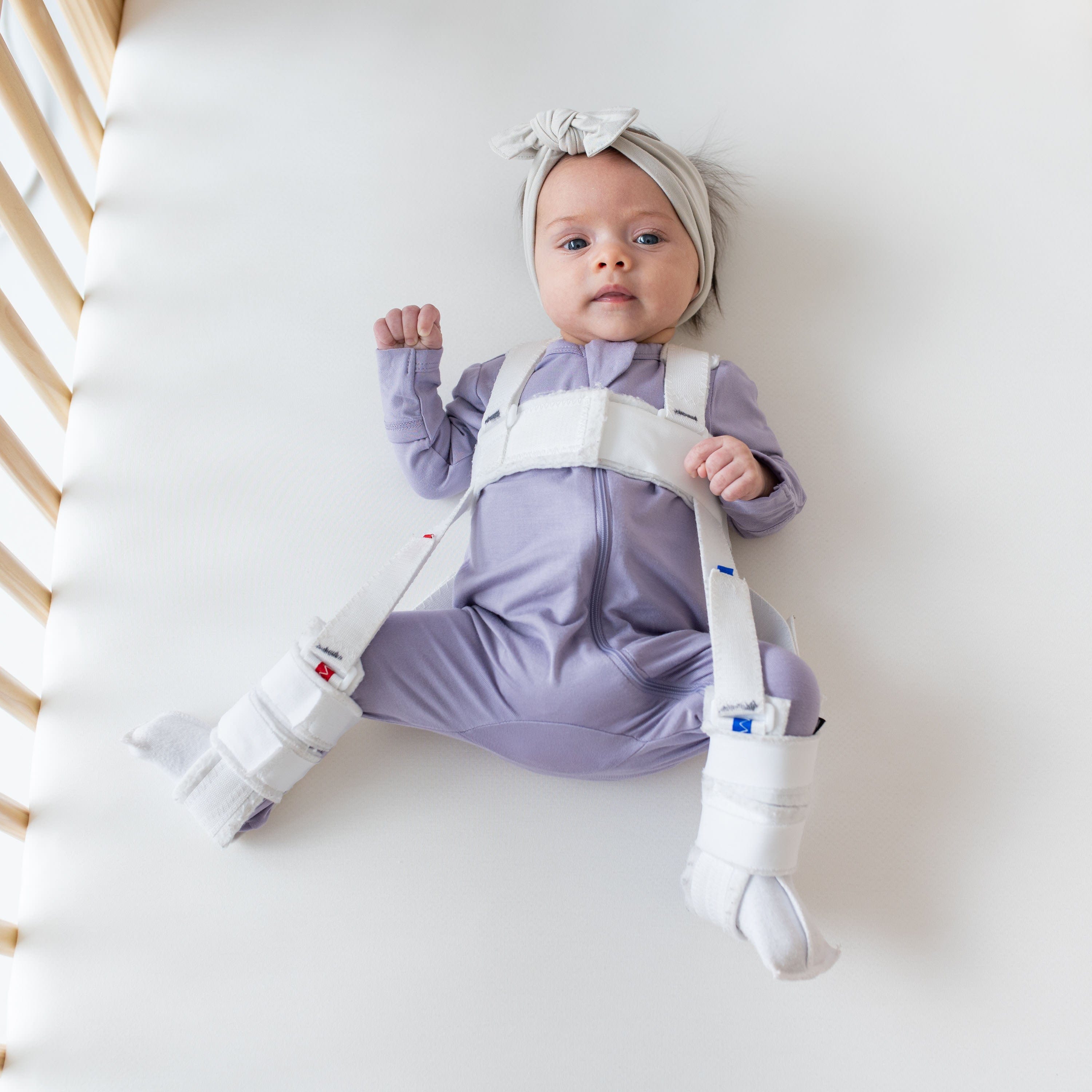 Baby laying in a crib while wearing a Zippered Romper in Taro and headband bow in Oat