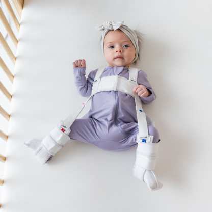 Baby laying in a crib while wearing a Zippered Romper in Taro and headband bow in Oat