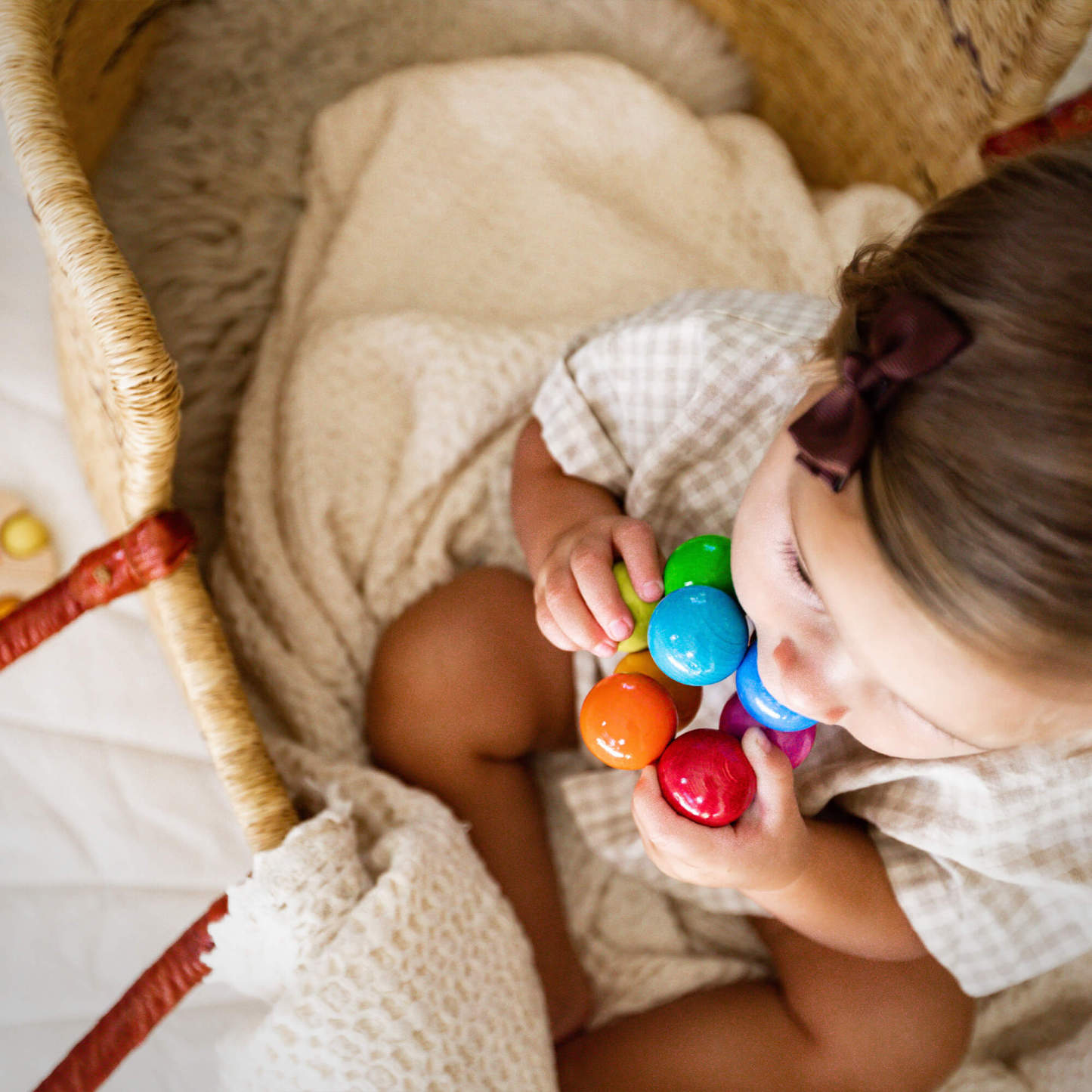 A young child with a bow in her hair sits in a basket, holding a Magica Wooden Clutching Toy while wrapped in a cozy blanket.