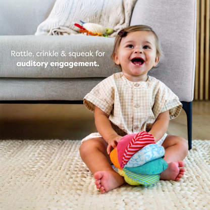 A young child sitting on a rug with a colorful toy ball, in front of a sofa with a blanket and toys. Text reads 'Rattle, crinkle & squeak for auditory engagement."