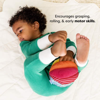A child in a green outfit lies on a white quilt, holding a colorful stuffed ball. Text reads "Encourages grasping, rolling, & early motor skills."