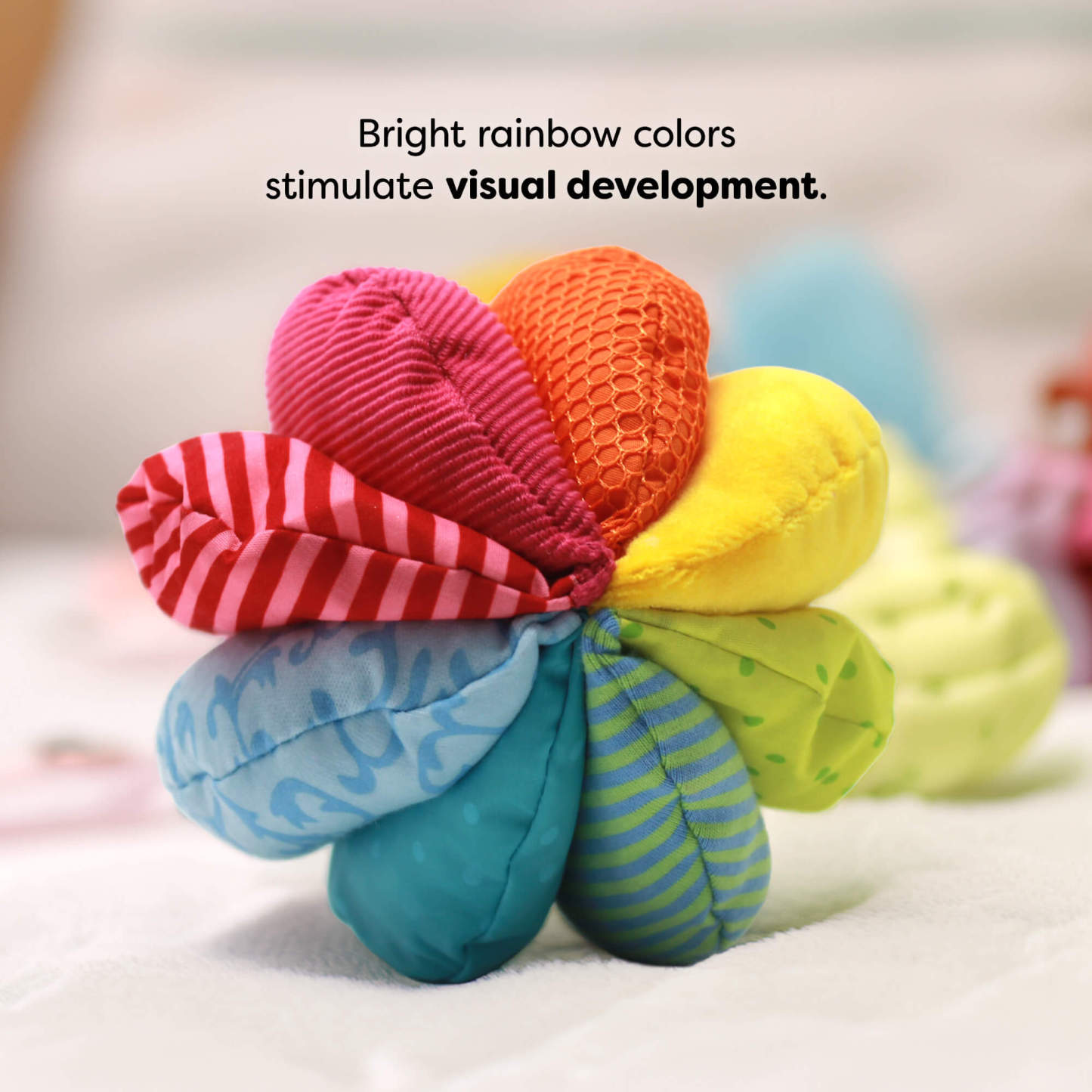 Colorful fabric toy with textured petals on a soft surface. Text reads "Bright rainbow colors stimulate visual development."