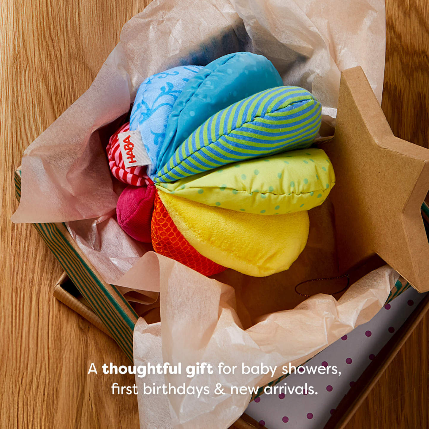 A colorful fabric ball inside an open gift box lined with tissue paper. Text reads "A thoughtful gift for baby showers, first birthdays & new arrivals."