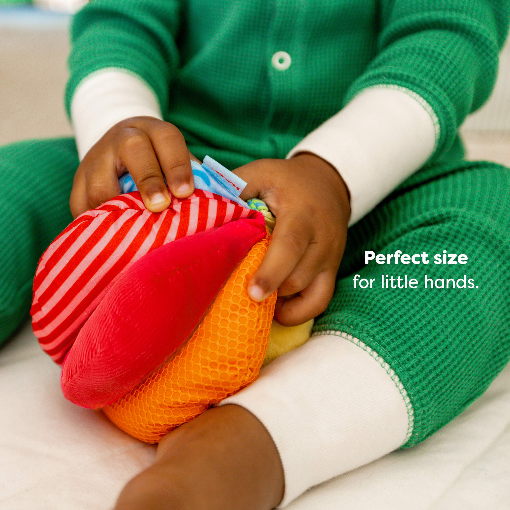 A child in green pajamas holding a colorful soft ball with striped and mesh textures. Text reads "Perfect size for little hands."