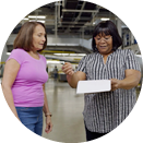 Two women are standing in a brightly lit industrial space. The woman on the left is wearing a pink shirt and jeans, while the woman on the right is in a striped shirt and dark pants, holding a clipboard and pointing at it. They appear to be engaged in conversation.