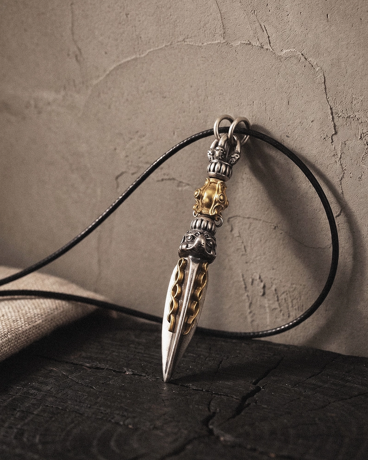 Phurba pendant necklace hanging from a black cord against a warm gray wall, with the pendant tip resting on dark wood.