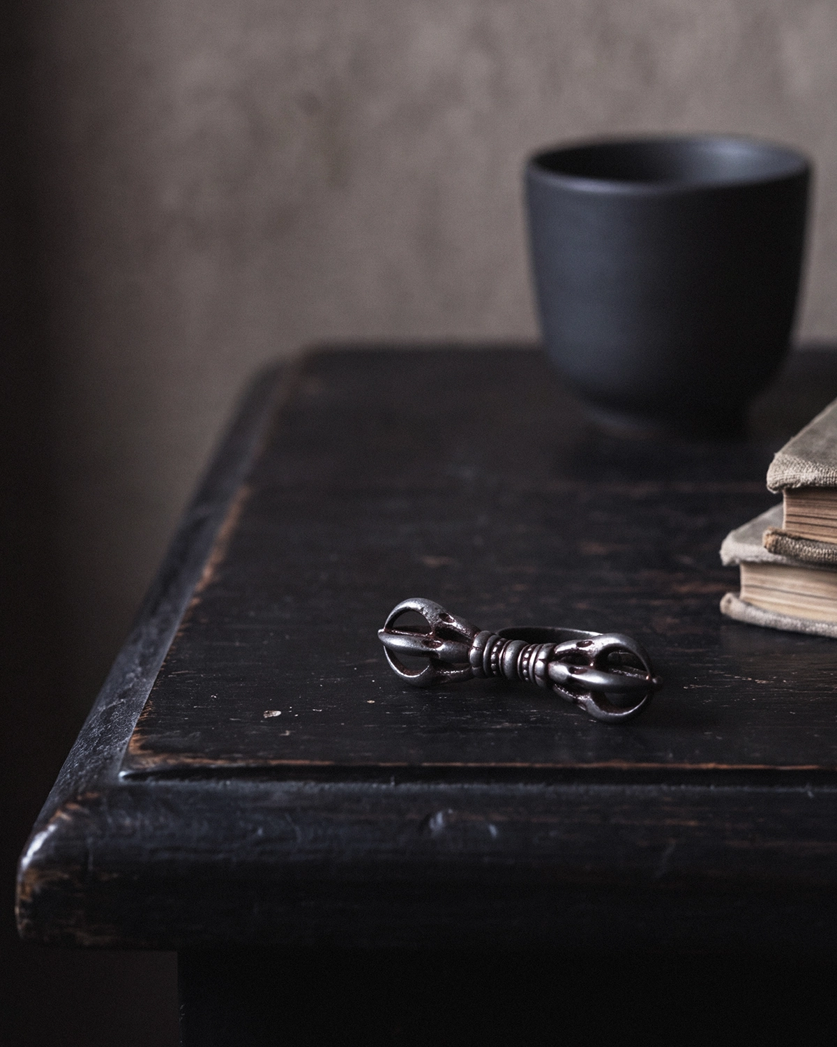 Vajra Dorje cold iron ring resting on a dark wood table with a black ceramic cup and stacked old books softly blurred behind it.