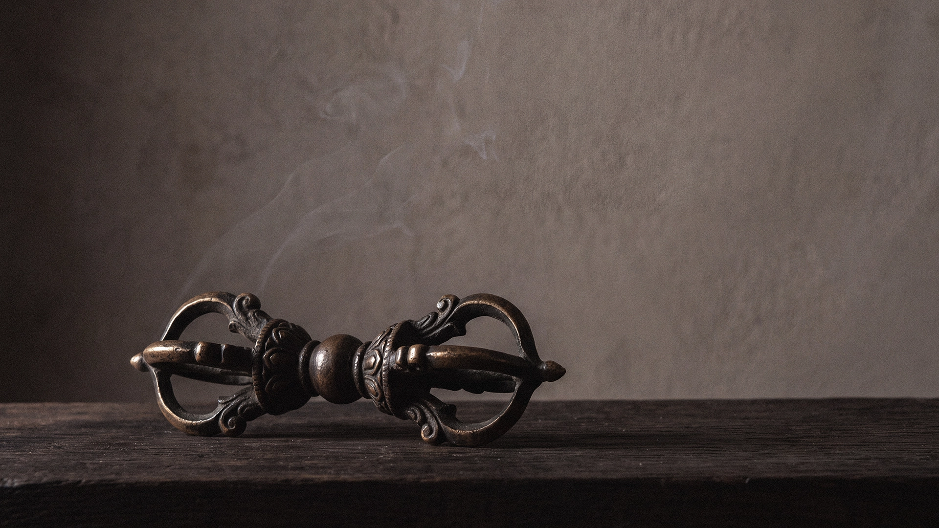 A Tibetan-inspired dorje, also called a vajra, resting on dark aged wood with a faint drift of incense smoke rising behind it.