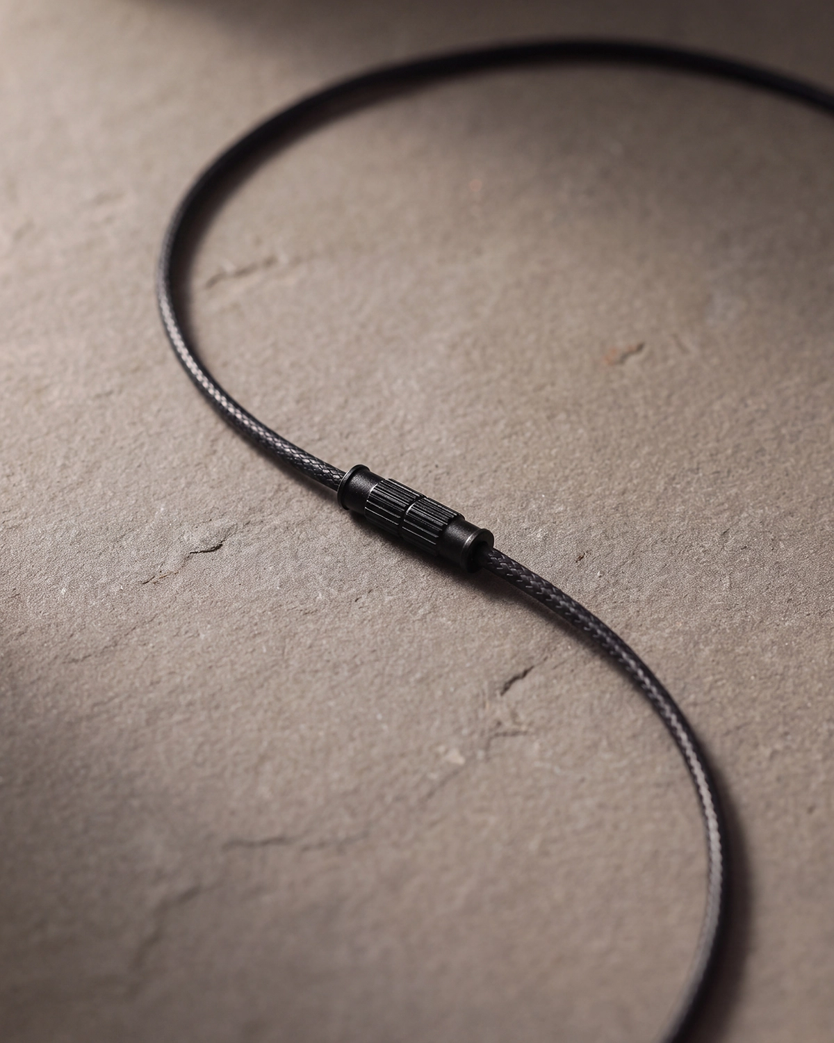 Detail of the black braided necklace cord with a black barrel screw clasp on a neutral stone surface.