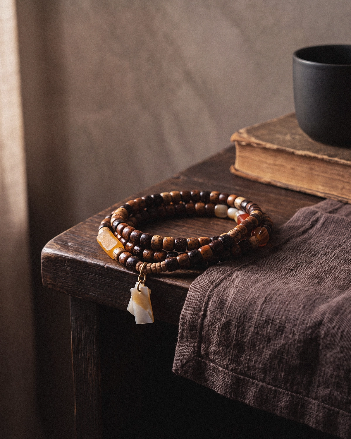 Star and moon bodhi seed mala resting on the edge of a dark wooden table beside brown linen, an old book, and a black cup.
