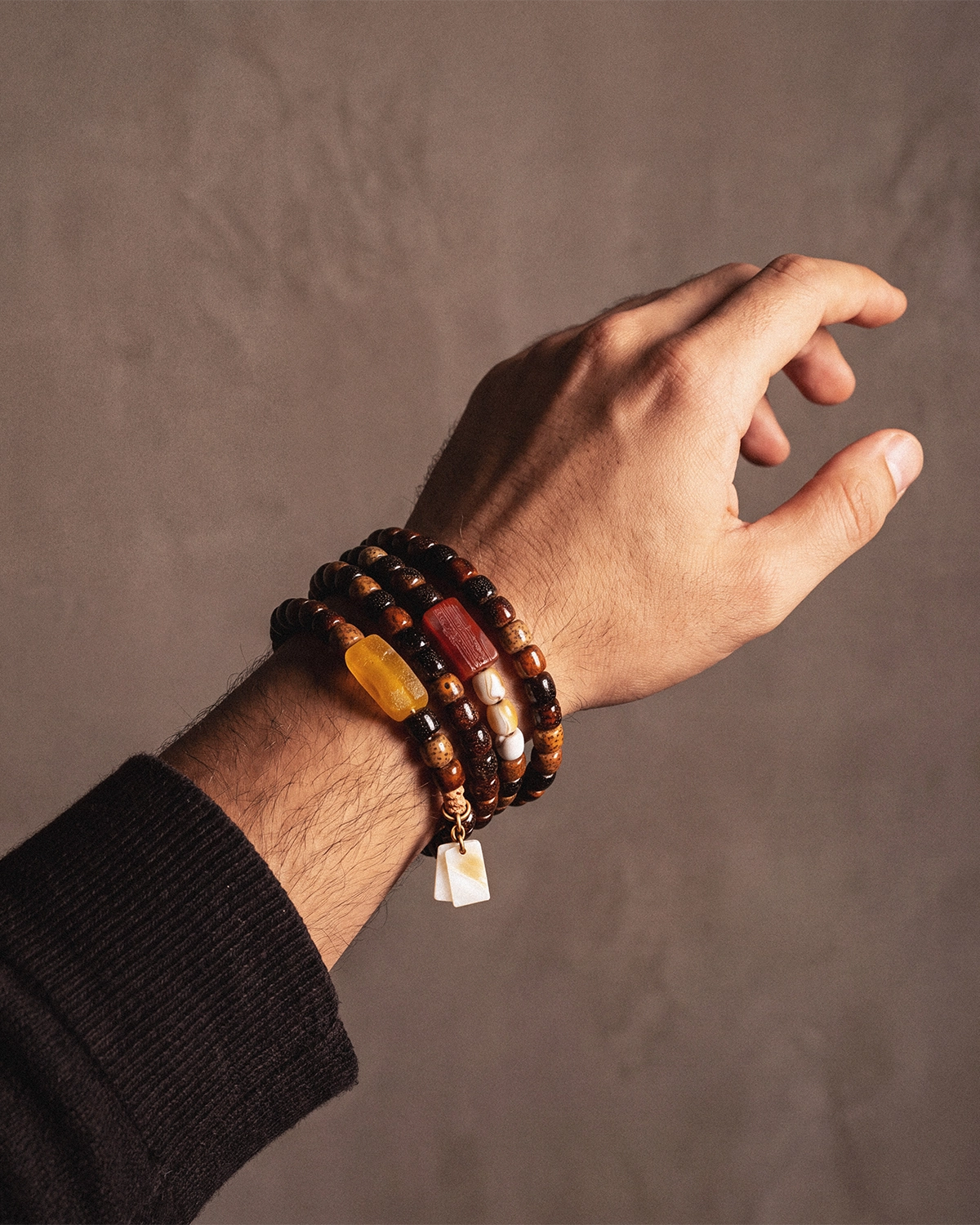 A 108-bead bodhi seed mala wrapped four times around a man's wrist with a black knit sleeve in view against a warm gray wall.