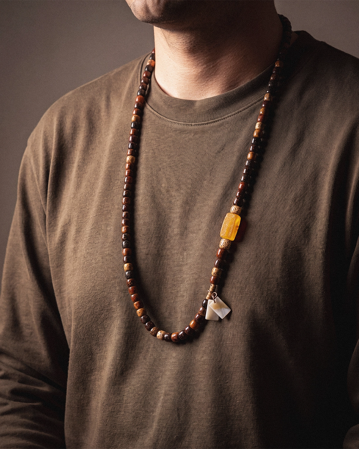 A star and moon bodhi seed mala worn long at a man's neckline over an olive-brown t-shirt, with the shell charm resting at the base of the strand.