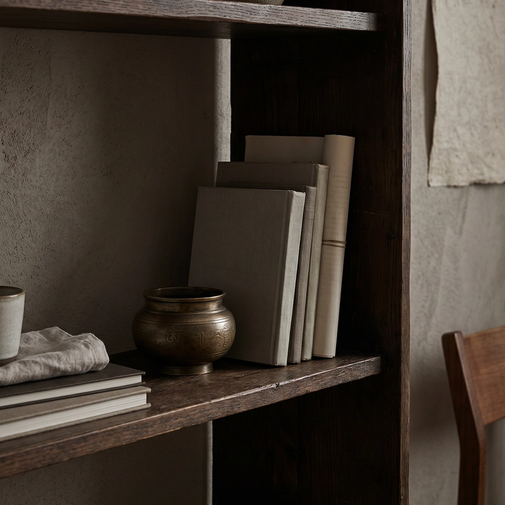 {"default":"Square mobile banner showing a quiet bookshelf scene with muted books, an aged metal vessel, ceramic cup, linen, dark wood, and restrained library styling."}
