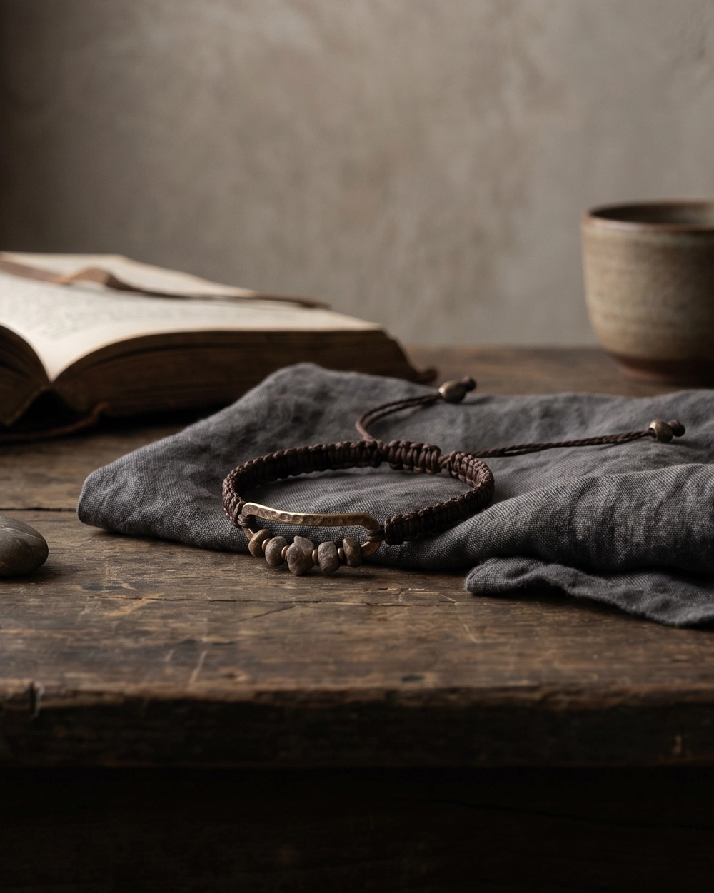 Handwoven bracelet on dark wood with linen, stones, an open book, and a ceramic cup in a quiet wabi-sabi interior