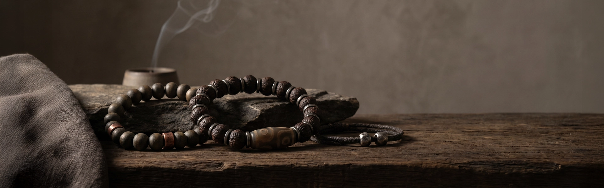 Dzi-style and bodhi bead bracelets arranged on raw stone and dark wood in a quiet Western wabi-sabi ritual setting with soft smoke and warm shadow.