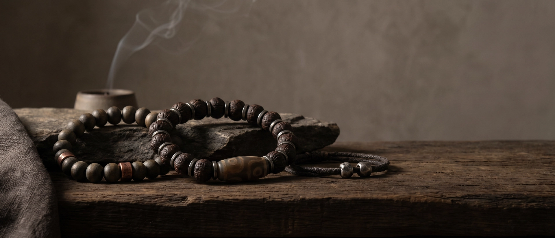 Dzi-style and bodhi bead bracelets arranged on raw stone and dark wood in a quiet Western wabi-sabi ritual setting with soft smoke and warm shadow.