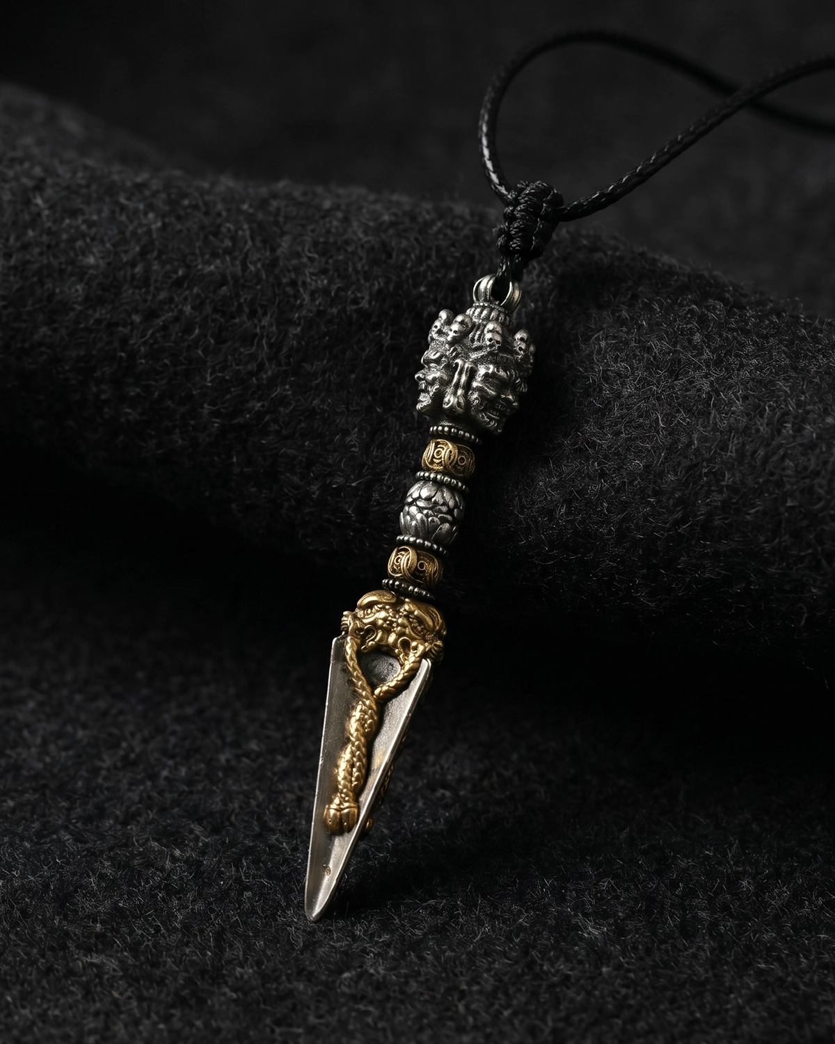 Brass Phurba pendant necklace with a black cord resting on dark textured fabric in a moody product-focused view.