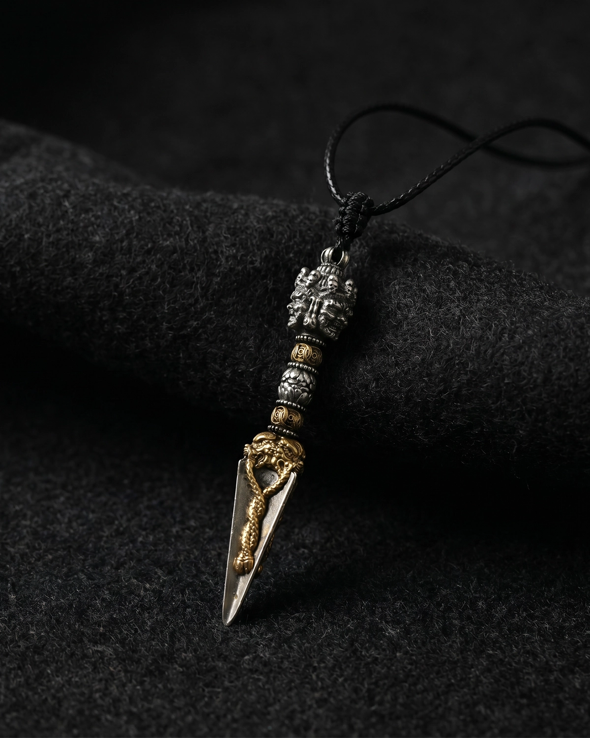 Brass Phurba pendant necklace with a black cord resting on dark textured fabric in a moody product-focused view.