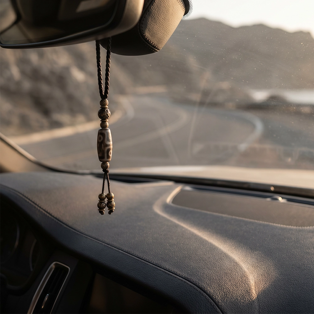 {"default":"Square mobile banner showing a Tibetan-inspired Dzi bead car charm hanging from a rearview mirror inside a quiet modern car interior."}

