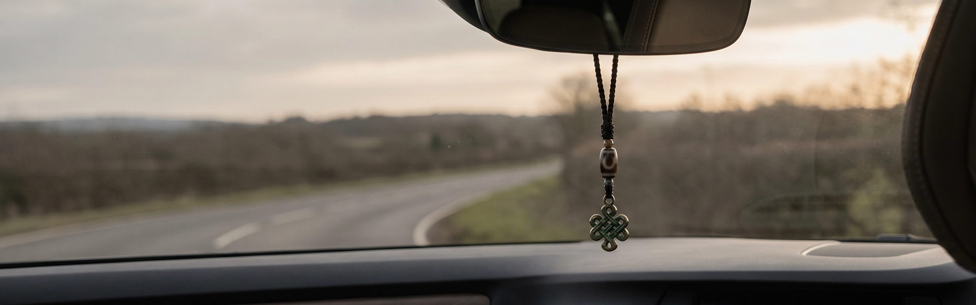 Tibetan-inspired car charm hanging from a rearview mirror with dark cord, muted beads, symbolic metal detail, and a quiet road travel view.