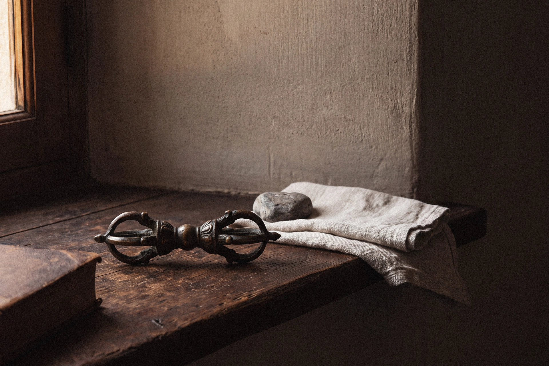 A dorje resting on a dark wooden desk near a window, beside the corner of an old book, a small gray stone, and folded linen cloth.