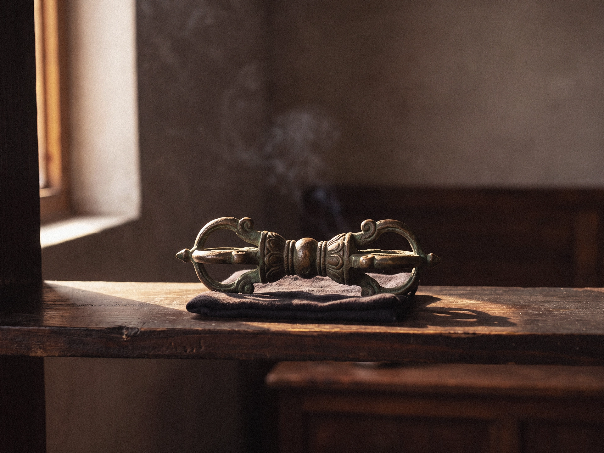 A metal double dorje with ornamental ends on both sides, resting on folded dark cloth on a wooden shelf near a sunlit window.