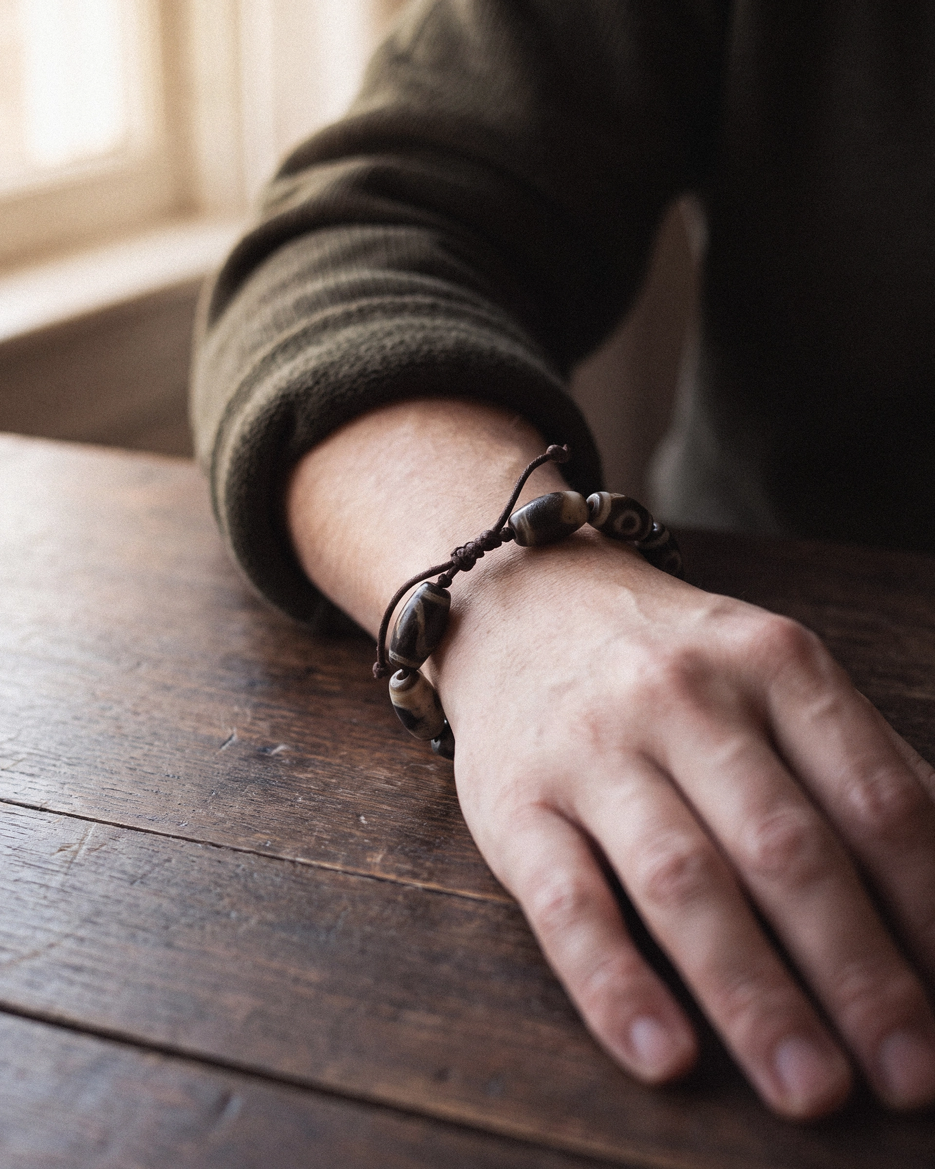 A person's wrist wearing a dzi bead bracelet on a dark cord, hand resting loosely on a wooden table.