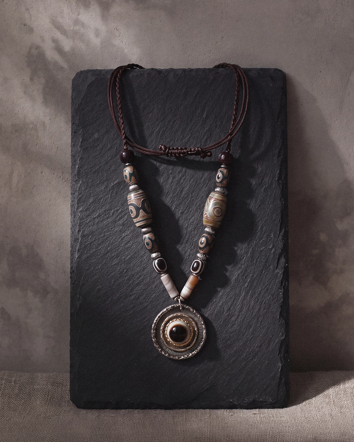 Dzi bead necklace with an eye agate pendant arranged on a dark slate slab against a warm gray wall.