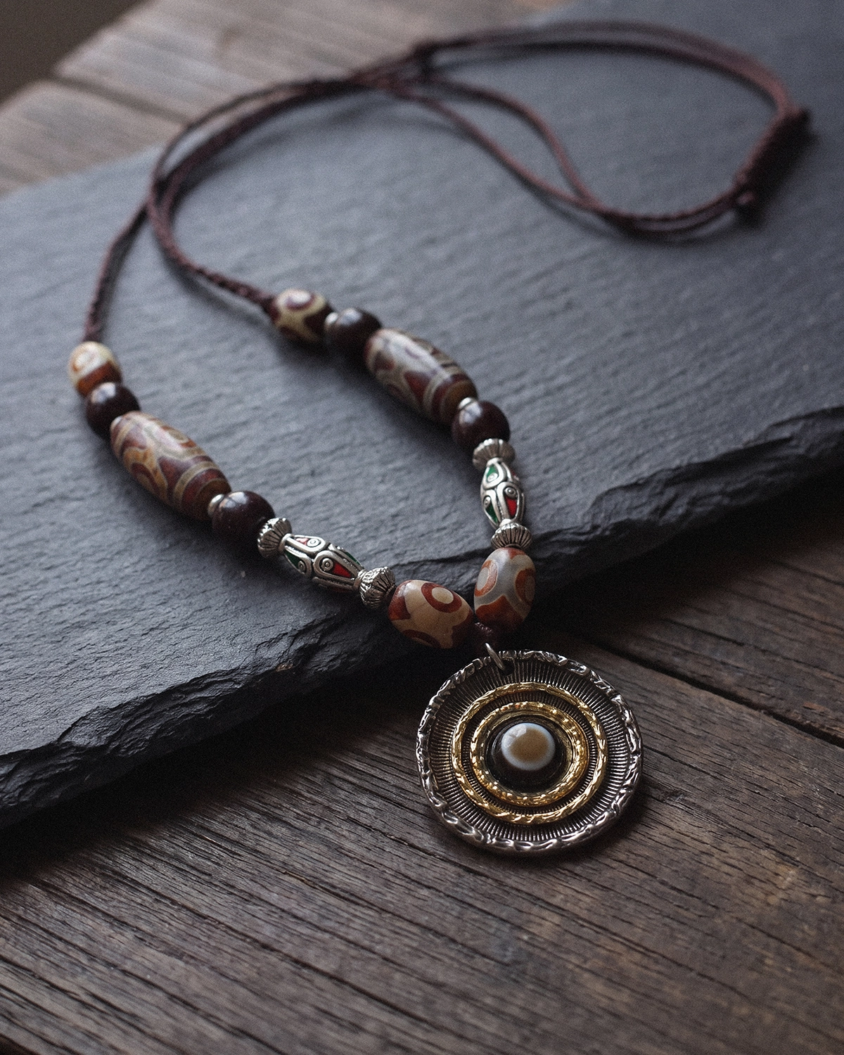 Dzi bead necklace with red-banded agate beads and a silver-toned eye pendant laid across dark slate on a wooden surface.