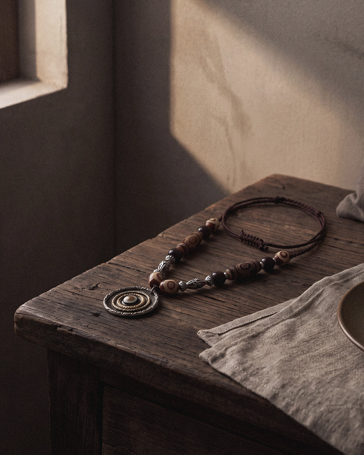 Dzi bead necklace resting on a weathered wooden table beside folded linen in quiet window light.