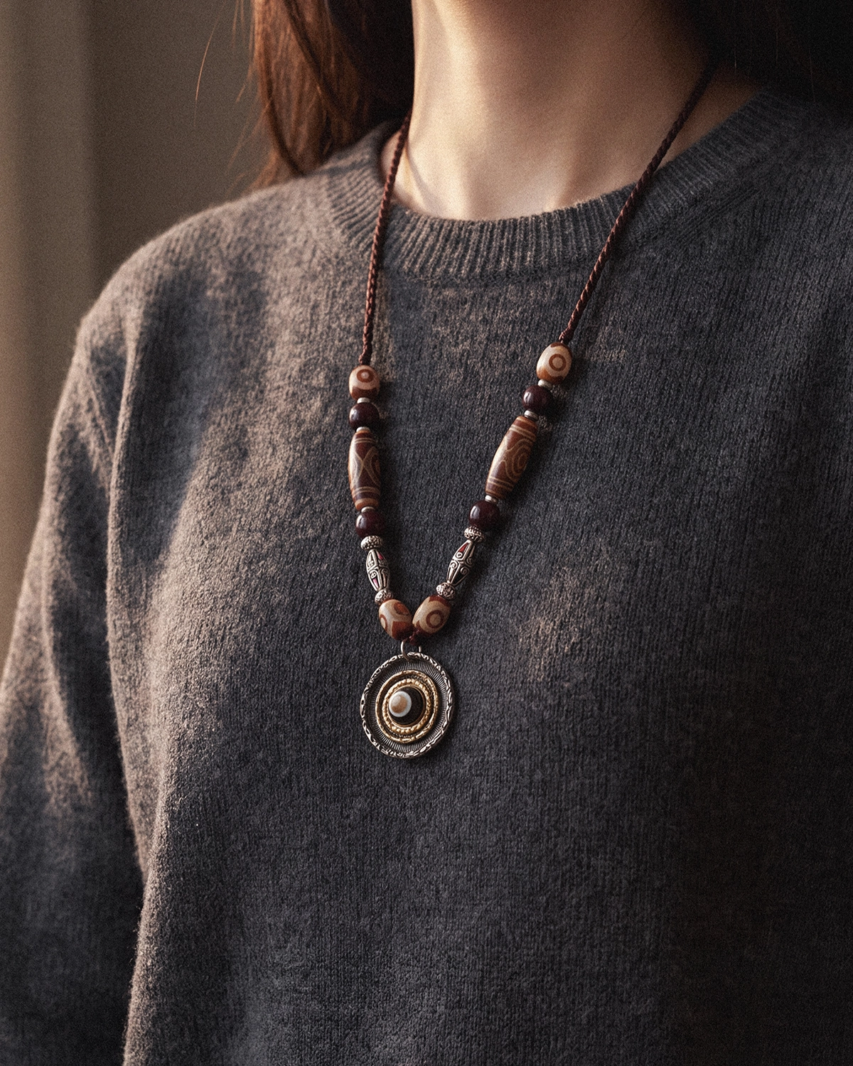 Warm-toned dzi bead necklace with a round eye pendant worn over a gray knit sweater.