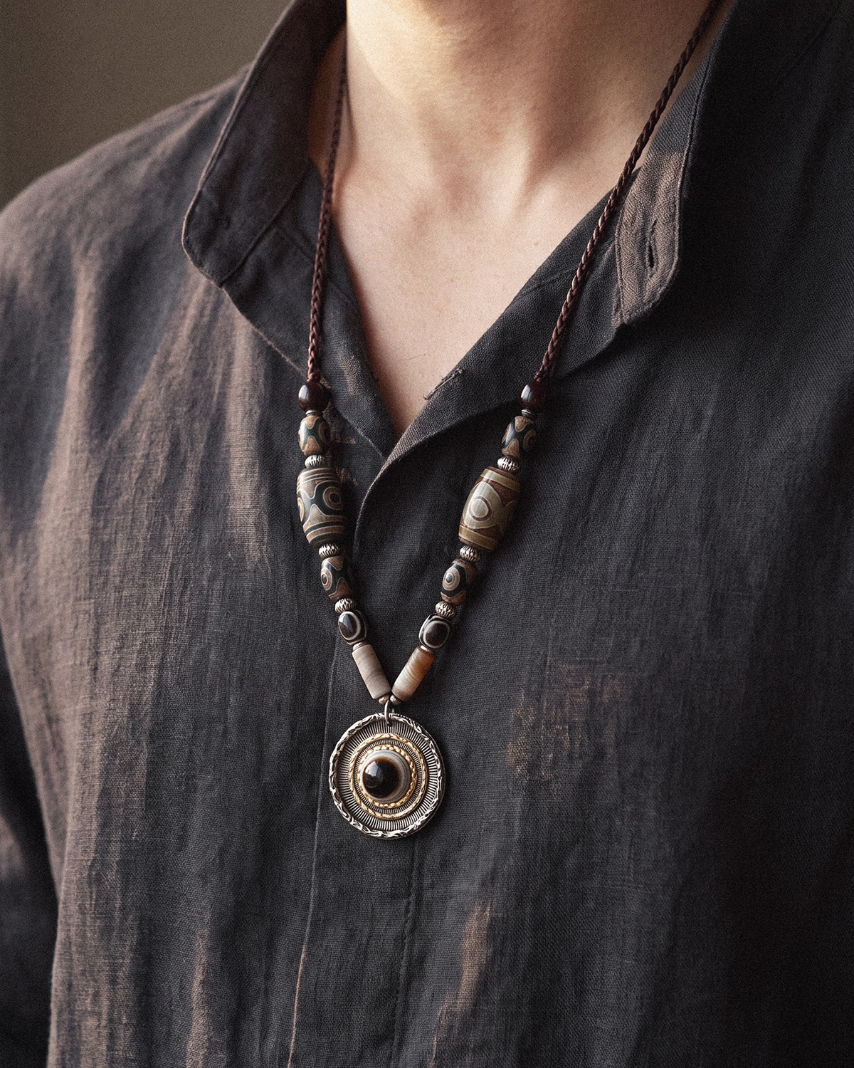 Dzi bead necklace worn at the collarbone over an open dark gray linen shirt, with the round eye pendant resting on the chest.