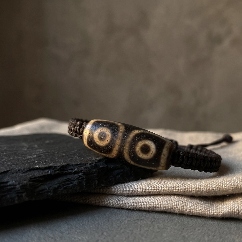 {"default":"Square mobile banner showing a Dzi-inspired bead cord bracelet on black slate and neutral linen in a quiet wabi-sabi setting."}
