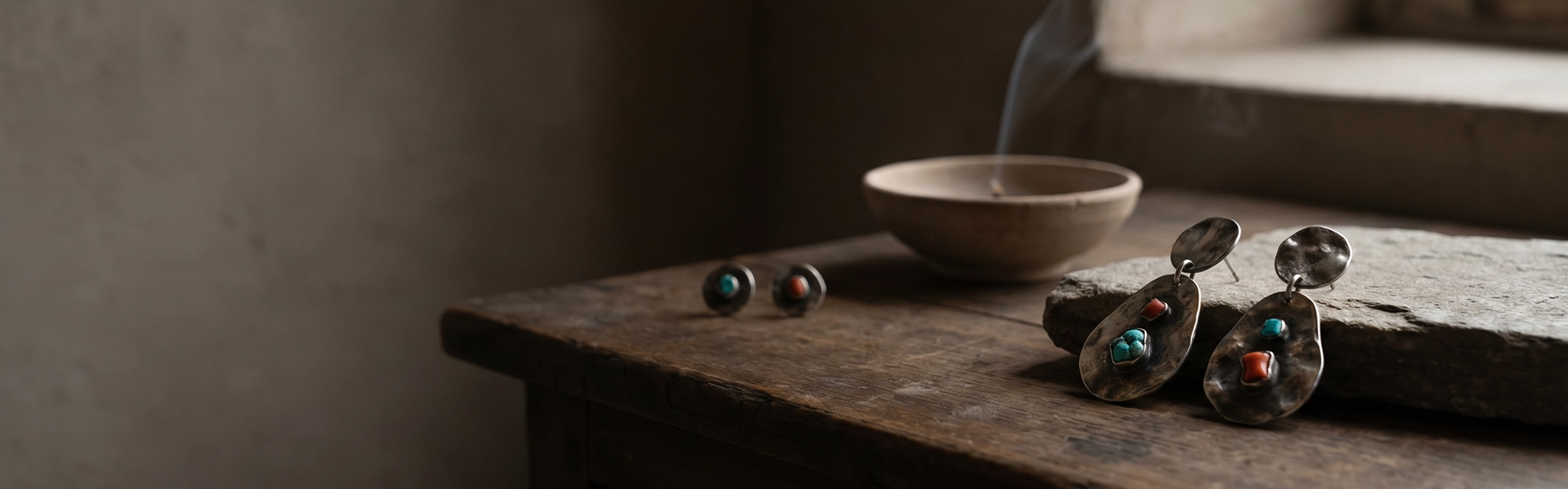Handcrafted Tibetan-inspired earrings with aged metal, turquoise and coral accents arranged on dark wood and raw stone in a quiet Western wabi-sabi ritual setting.