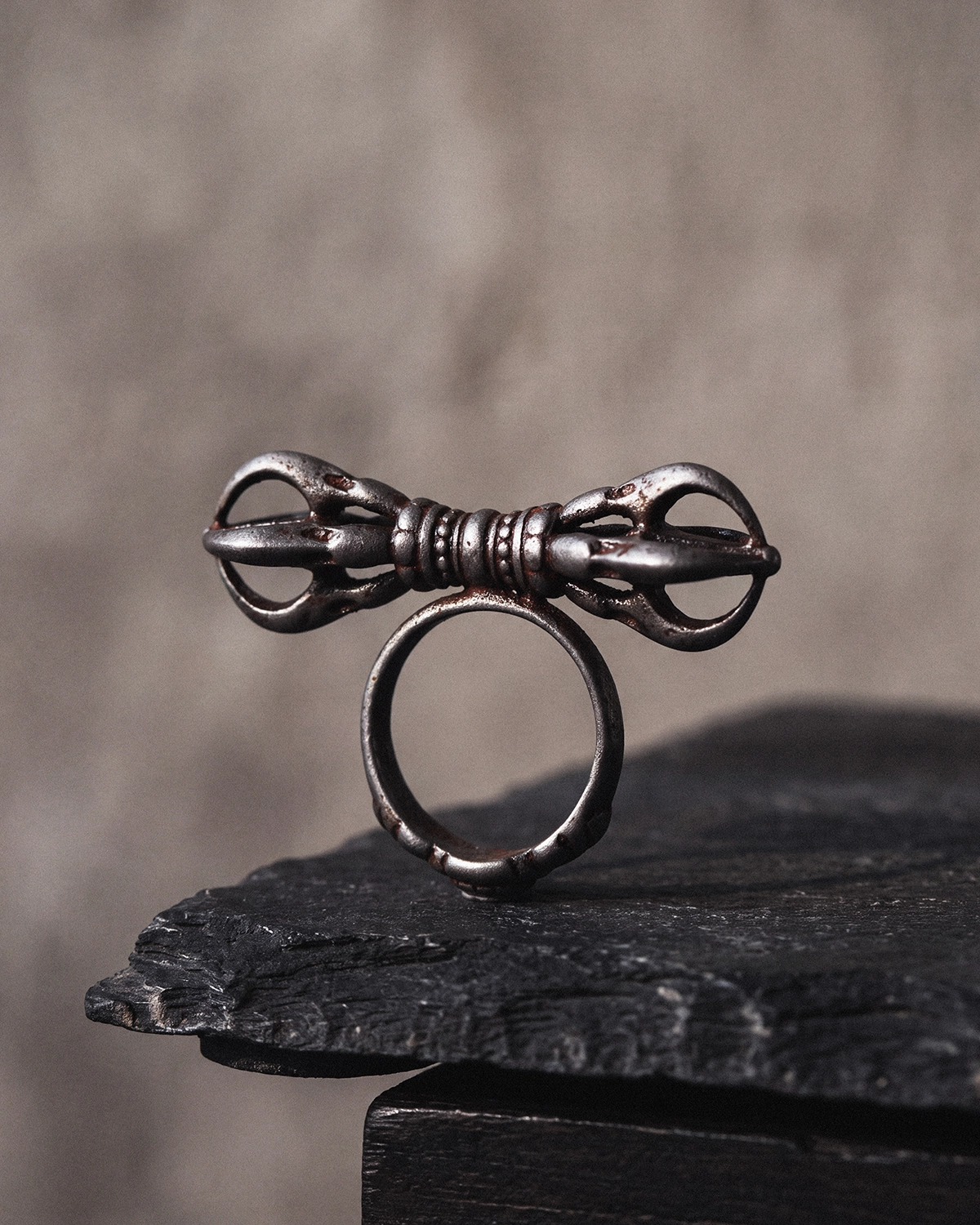 Cold iron Vajra Dorje ring standing upright on a dark stone slab against a warm gray wall.