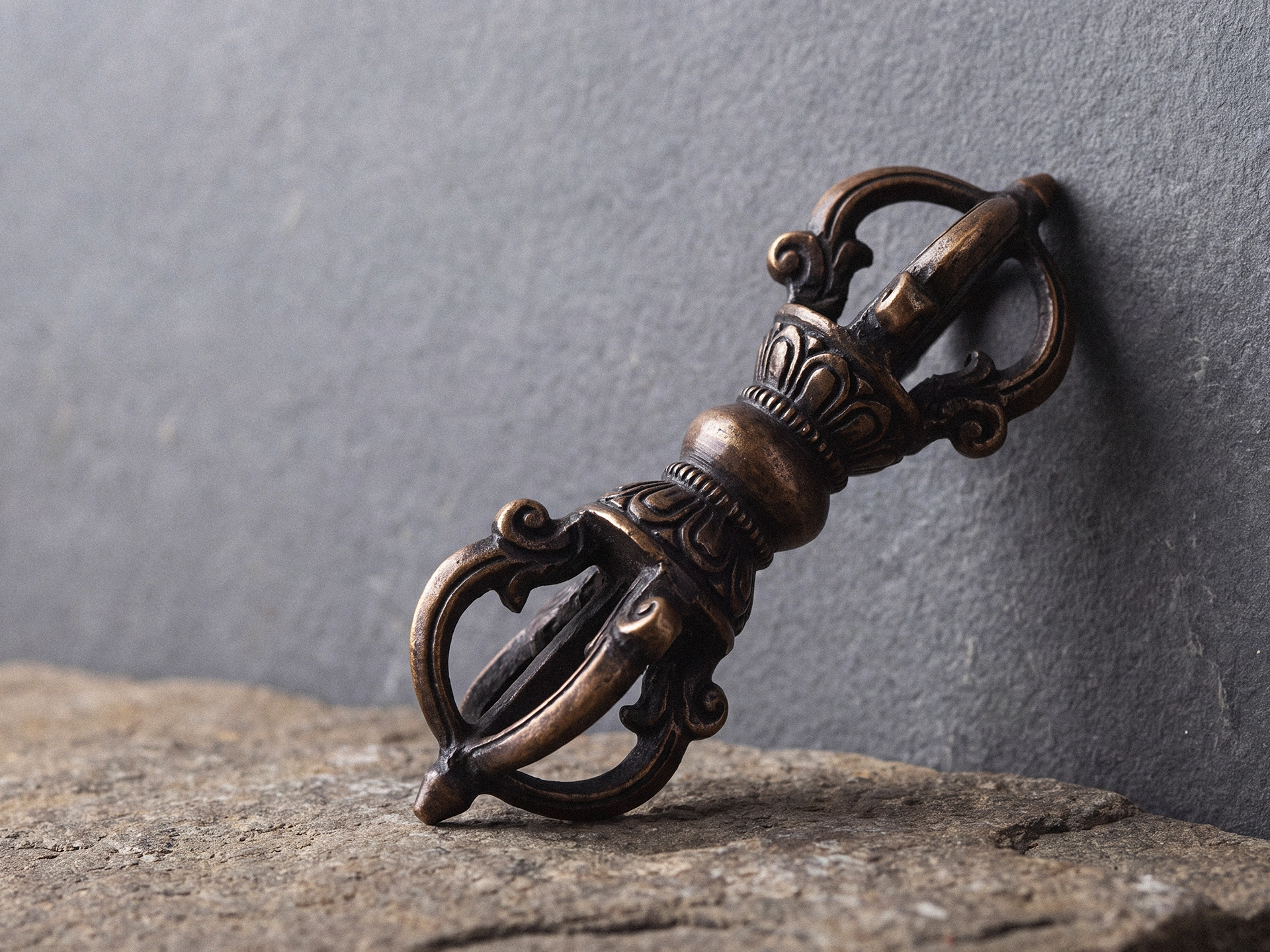 A three-quarter close-up of an aged bronze dorje showing its central grip, carved shaft, and curved prongs separating at each end.